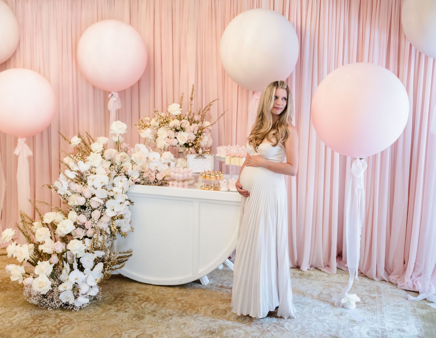Baby shower setup with white balloons, floral arrangements and pink drapery backdrop.