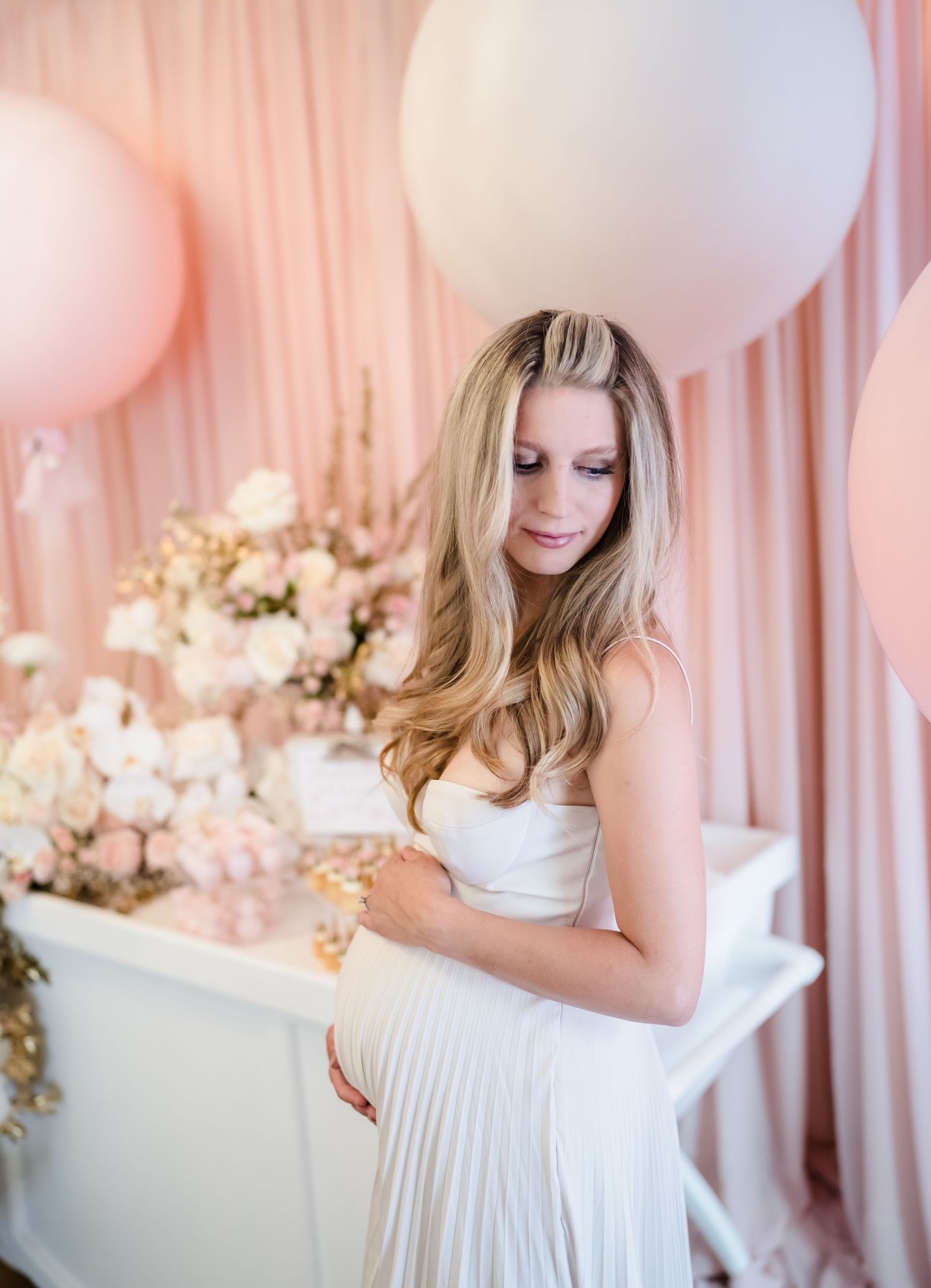 Maternity photo featuring elegant white gown against pink backdrop with balloons.