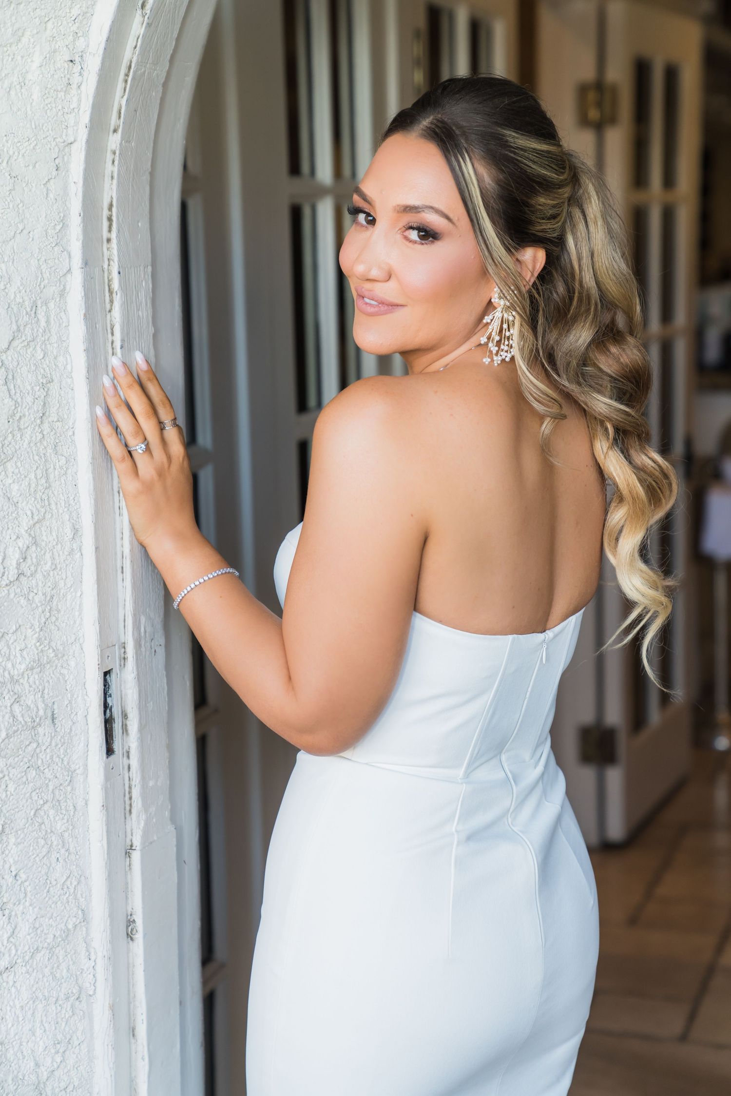 Elegant bridal portrait showing a white strapless dress, wavy blonde hair, and sparkling statement earrings.