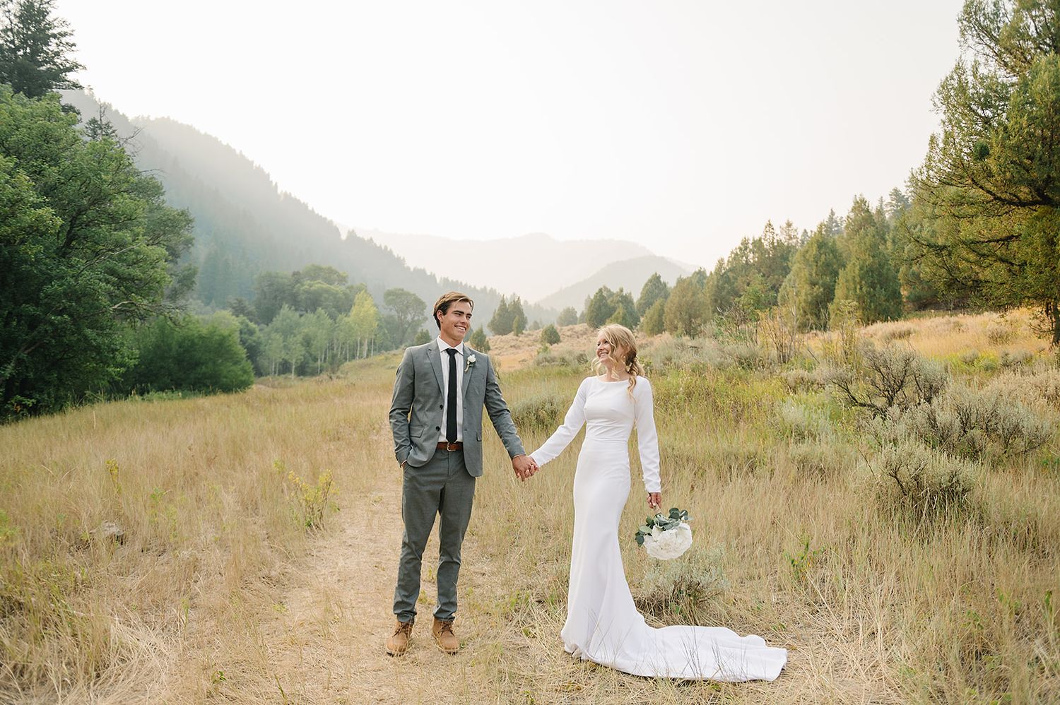 Newlyweds walking together holding hands in a mountain meadow with evergreen forest backdrop.