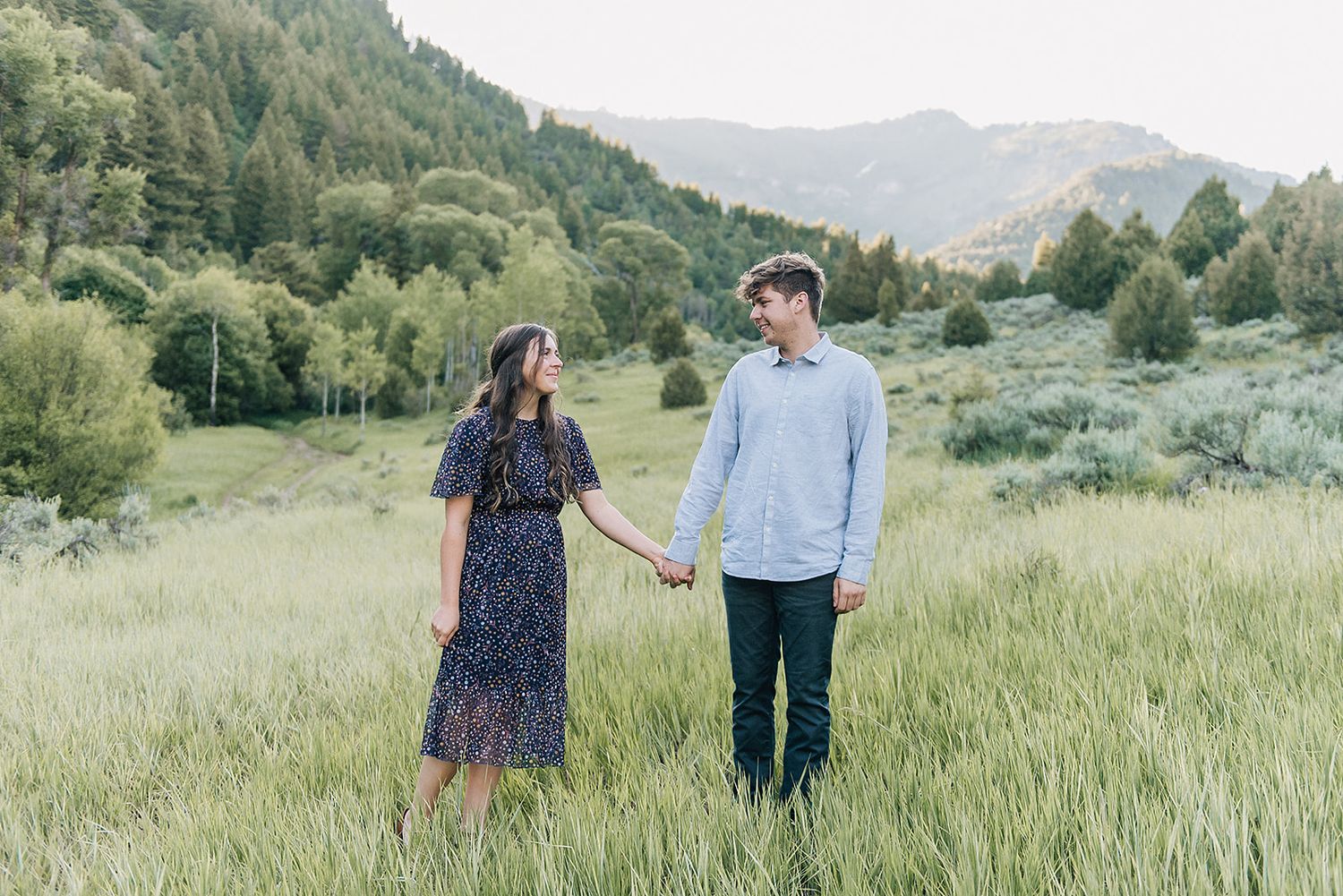 A couple poses in a mountain meadow holding hands with evergreen forests on the hillside.