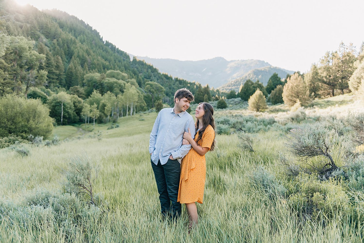 A couple stands close together in a mountain valley with sage brush and rolling hills.