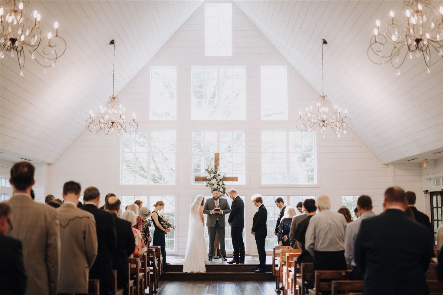 A wide view of a chapel wedding ceremony with guests seated in wooden pews.