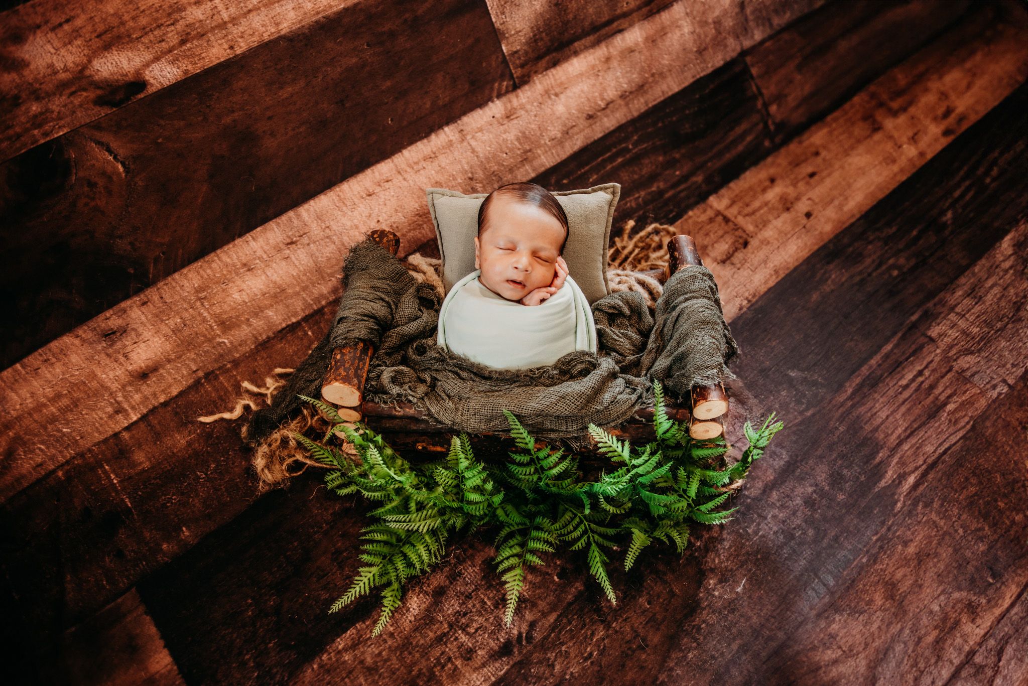 Sleeping newborn in basket surrounded by greenery on wooden floor.