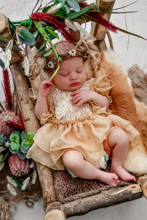Sleeping infant wearing peach-colored dress surrounded by floral decorations on rustic wooden surface.