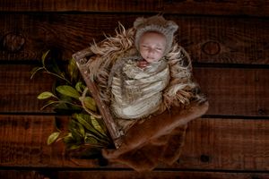 Newborn wrapped in beige fabric rests on rustic wooden backdrop with green foliage accents.