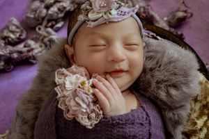 Sleeping baby in purple outfit with lace headband and fur wrap.
