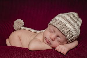 Sleeping baby wearing knit hat with pom pom lying on red fabric.