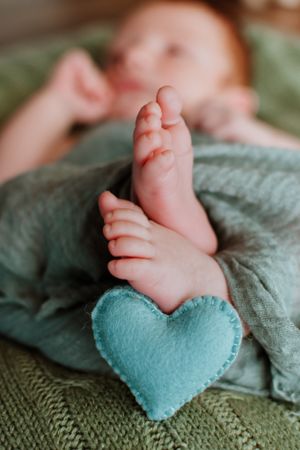 Tiny baby feet resting on teal heart-shaped pillow on knit blanket.