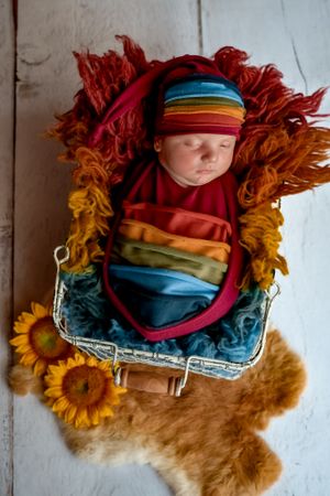 Newborn wrapped in rainbow colors nestled among sunflowers and colorful textiles.