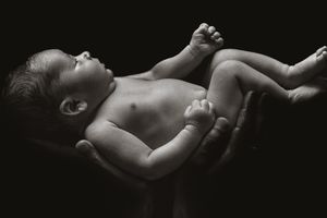 Artistic black and white portrait of infant lying on supportive hands in dramatic lighting.