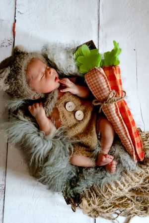 Sleeping baby in rustic outfit with decorative carrot prop on white wooden background.