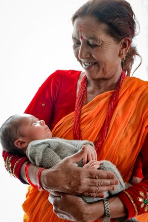 Woman in red and orange sari holding infant wrapped in grey sweater.