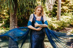 Woman in flowing blue dress holding baby in sunlit outdoor garden setting.