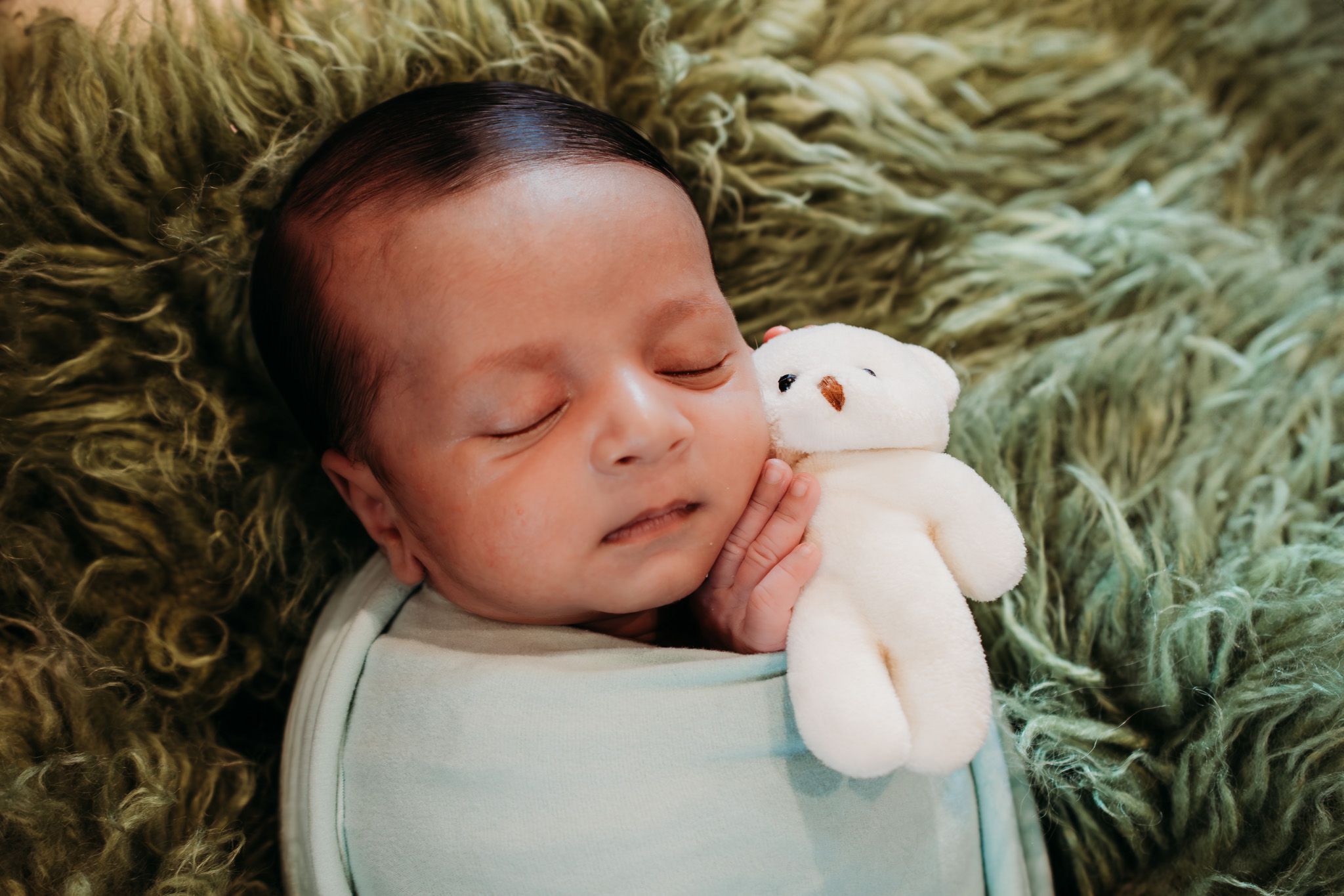 Peaceful sleeping infant on fuzzy green blanket with small white teddy bear.