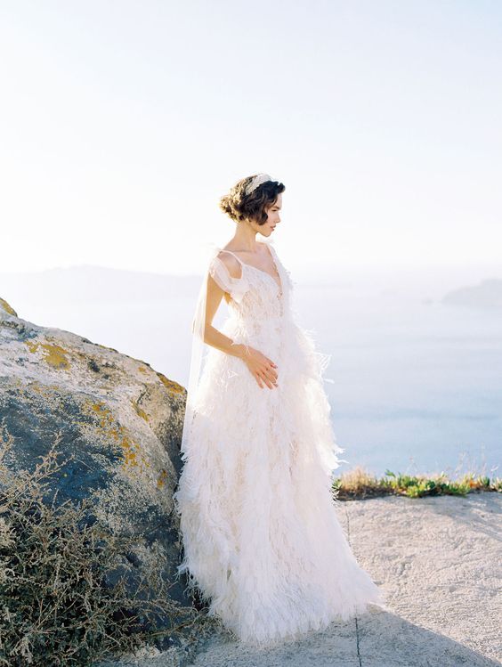 Ethereal bride in ruffled white gown poses dramatically on mountain cliff at sunset.