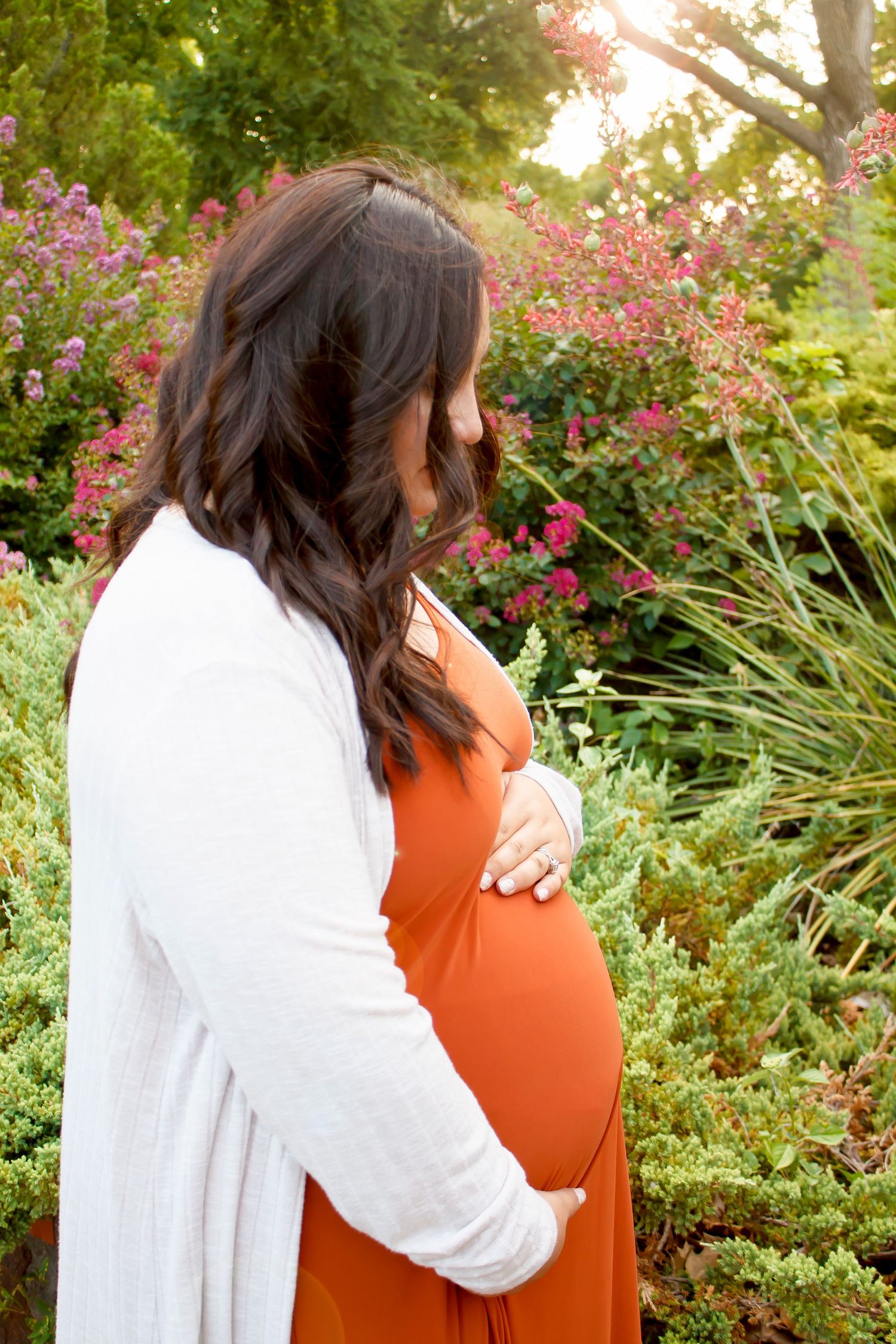 A maternity photo shows a woman in an orange dress and white cardigan standing among pink flowers in a garden.