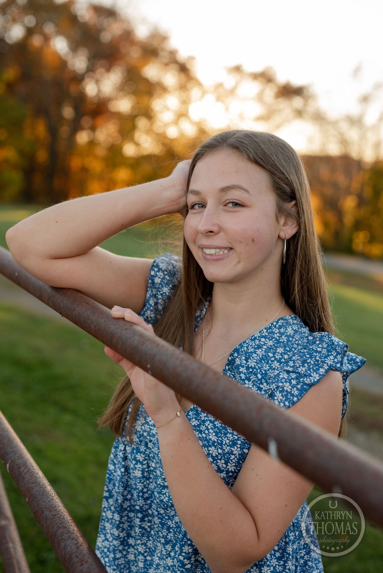 Someone poses casually against a fence rail during golden hour with fall foliage.