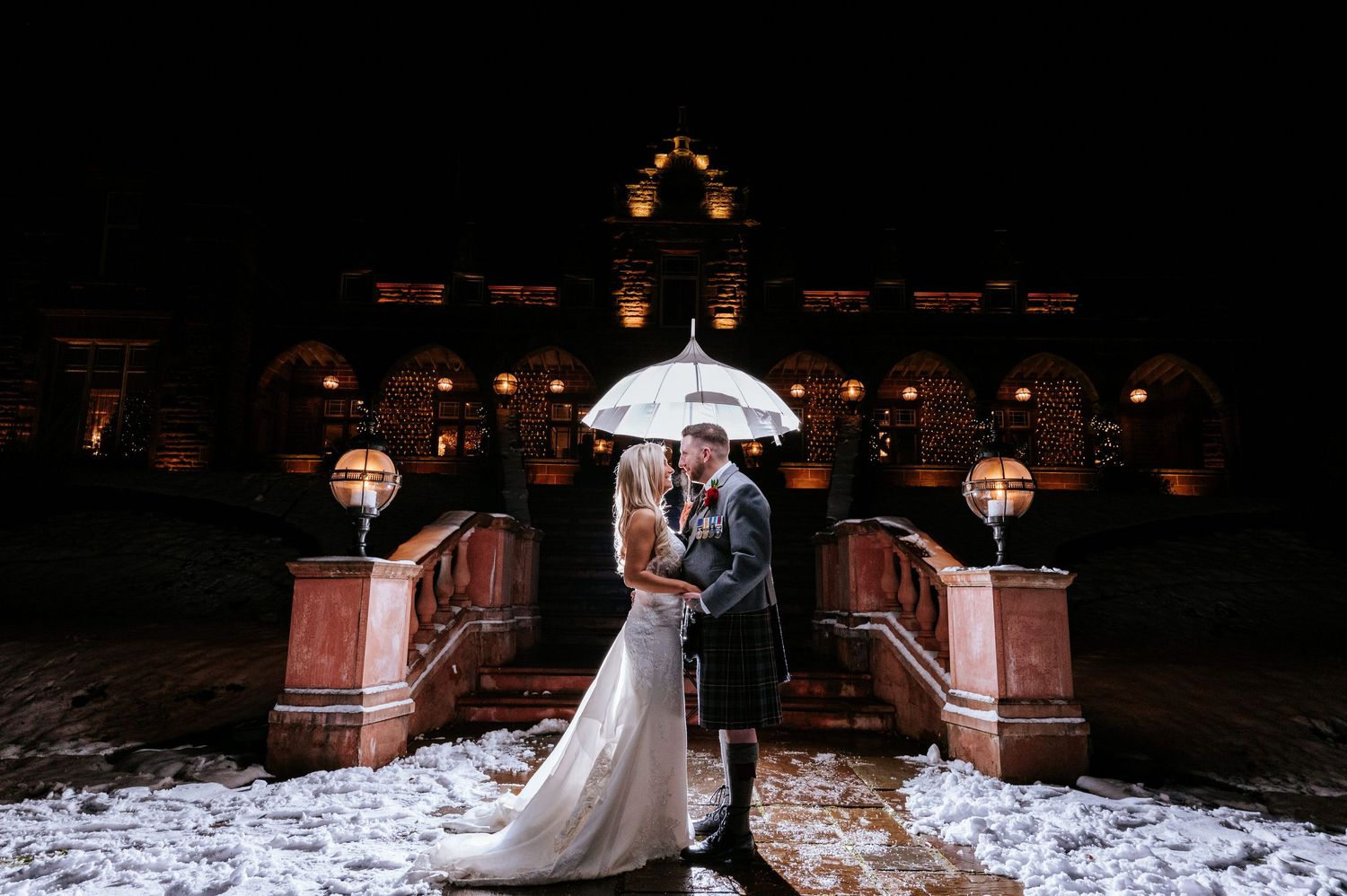 A winter wedding portrait at night with snow and an illuminated historic building in the background.