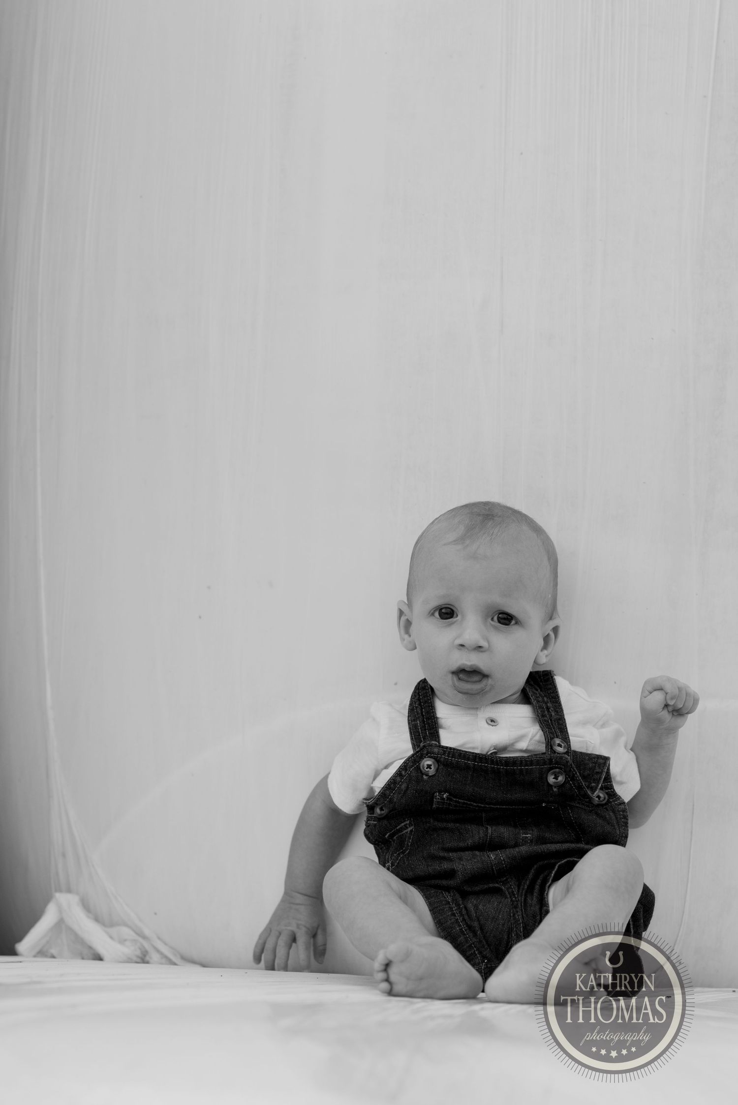 Black and white portrait of a baby wearing overalls sitting against a light background.