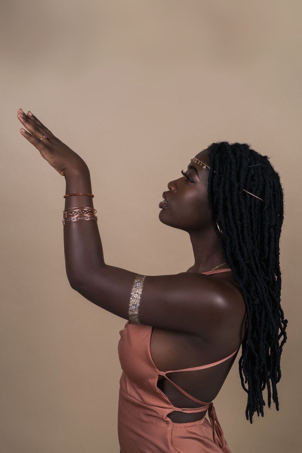 Artistic portrait of someone in coral dress with dreadlocks raising hand gracefully against neutral background.