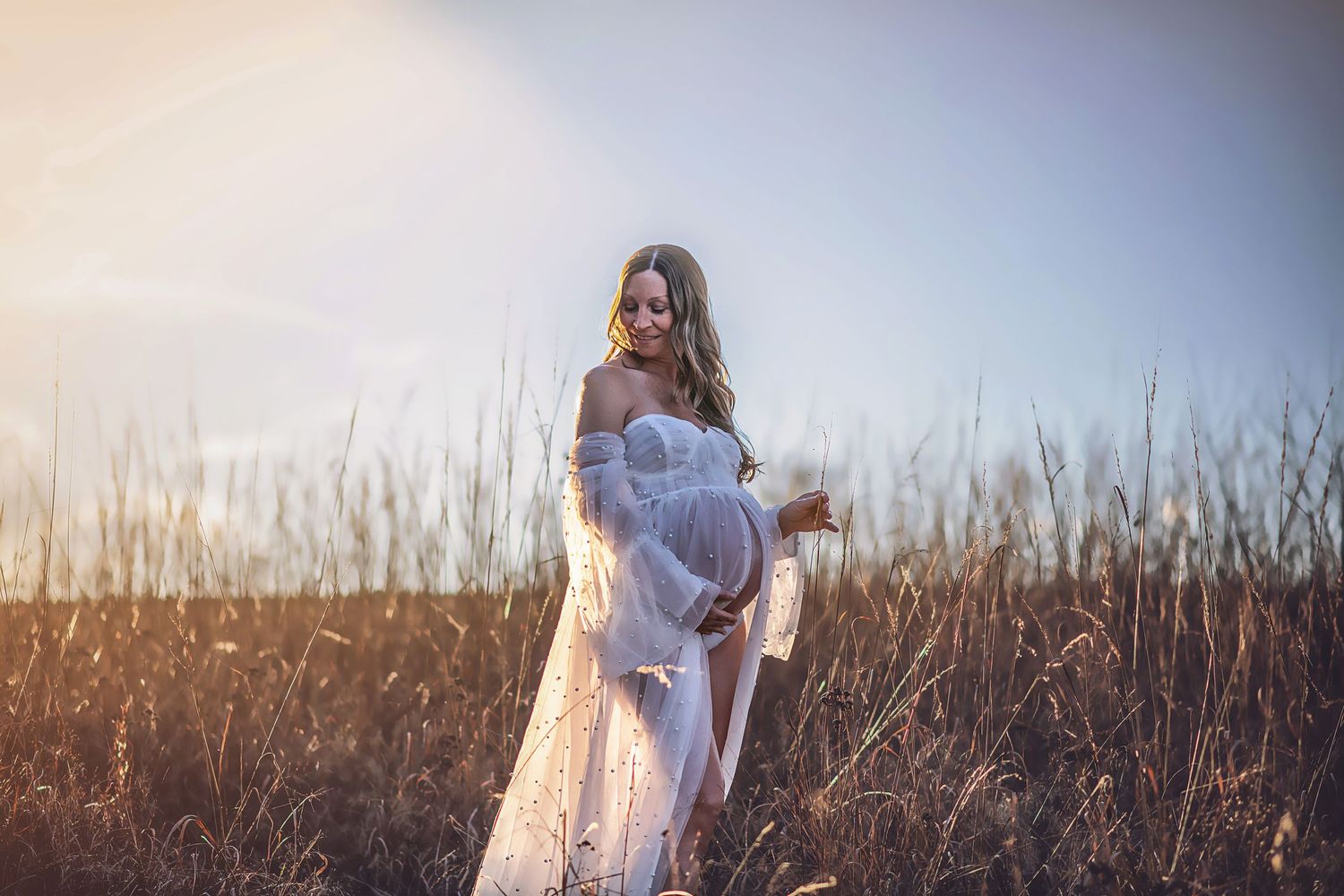 Ethereal maternity photo of woman in flowing dress among tall grass at sunset.