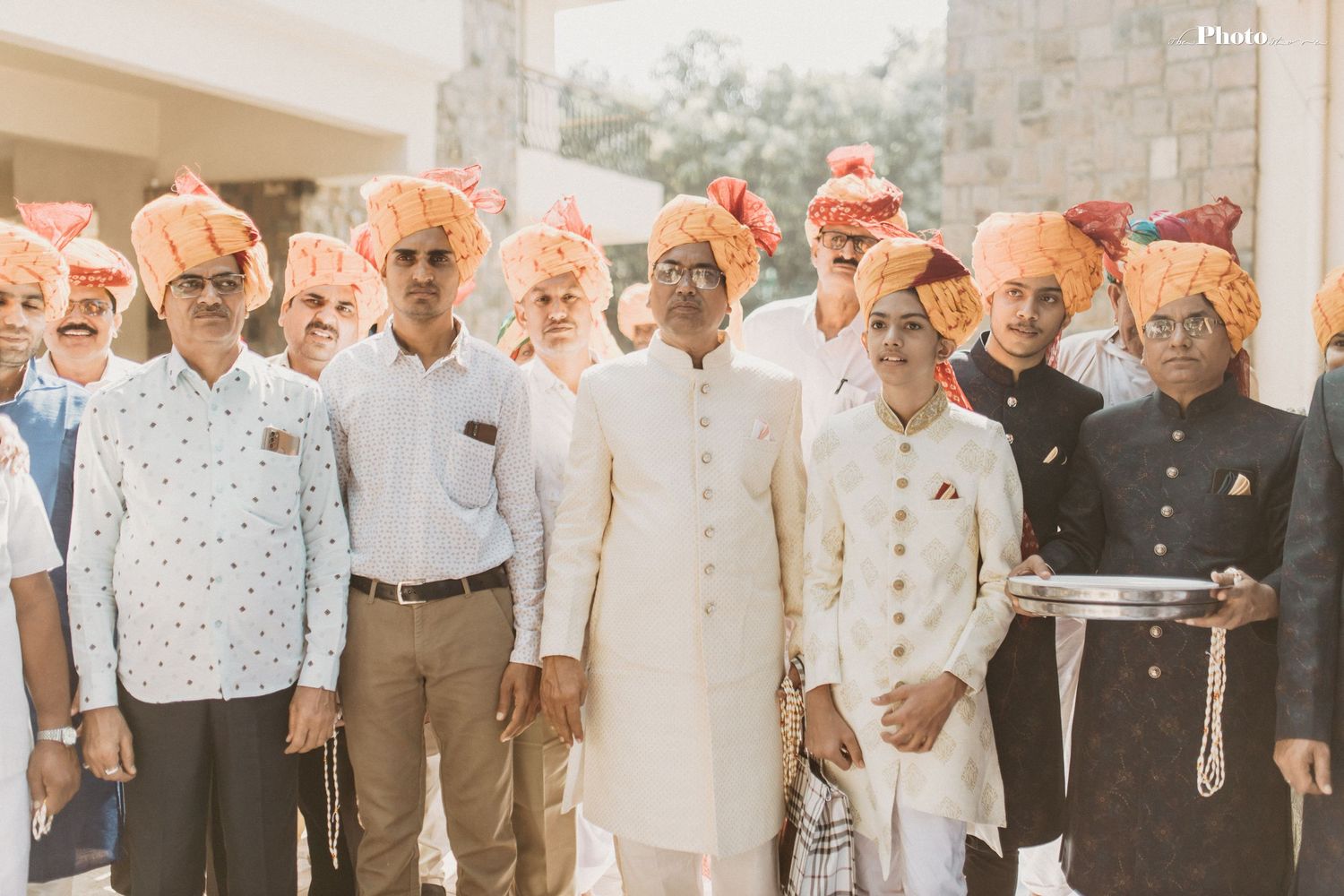 Group photo of wedding guests in traditional Indian formal attire against white building.