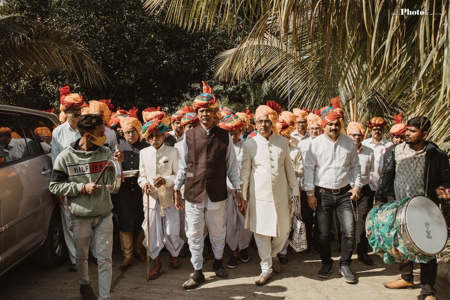 Large wedding party procession with guests in white and orange traditional attire under palm trees.