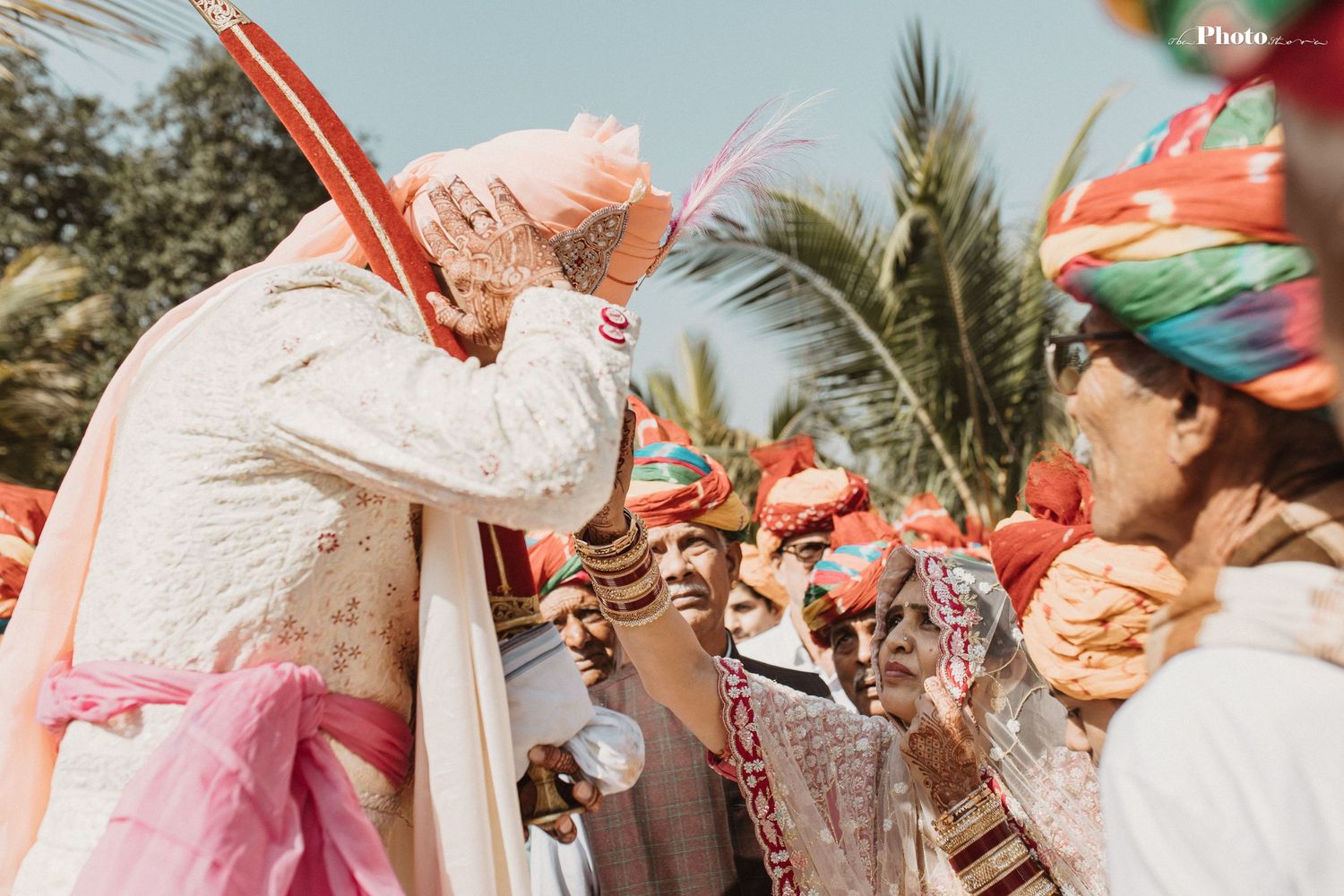 A joyous crowd celebrates at an outdoor Indian wedding ceremony with vibrant coral parasols and palm trees.