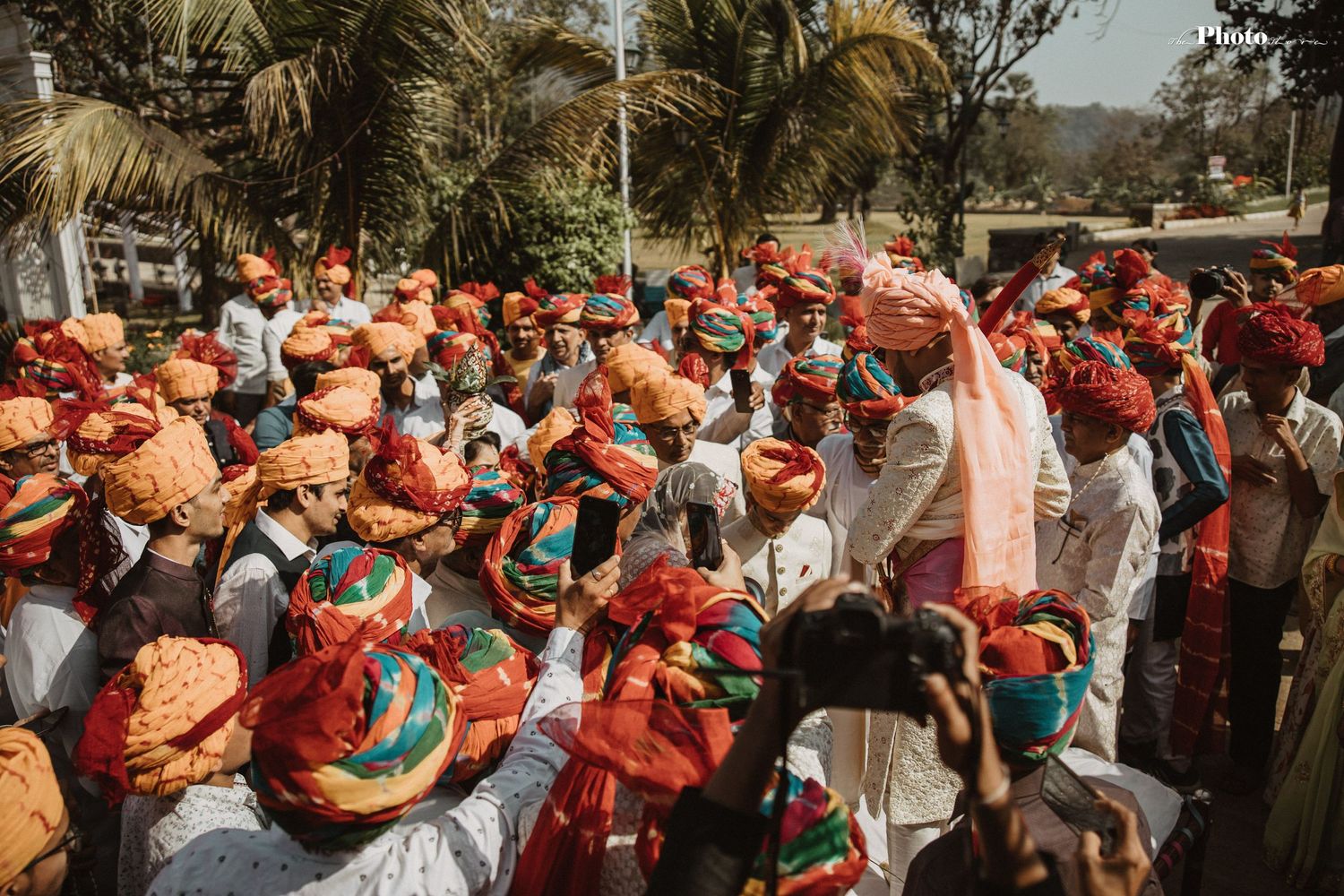 Festive crowd with coral umbrellas gathers under palm trees for traditional Indian wedding celebration.