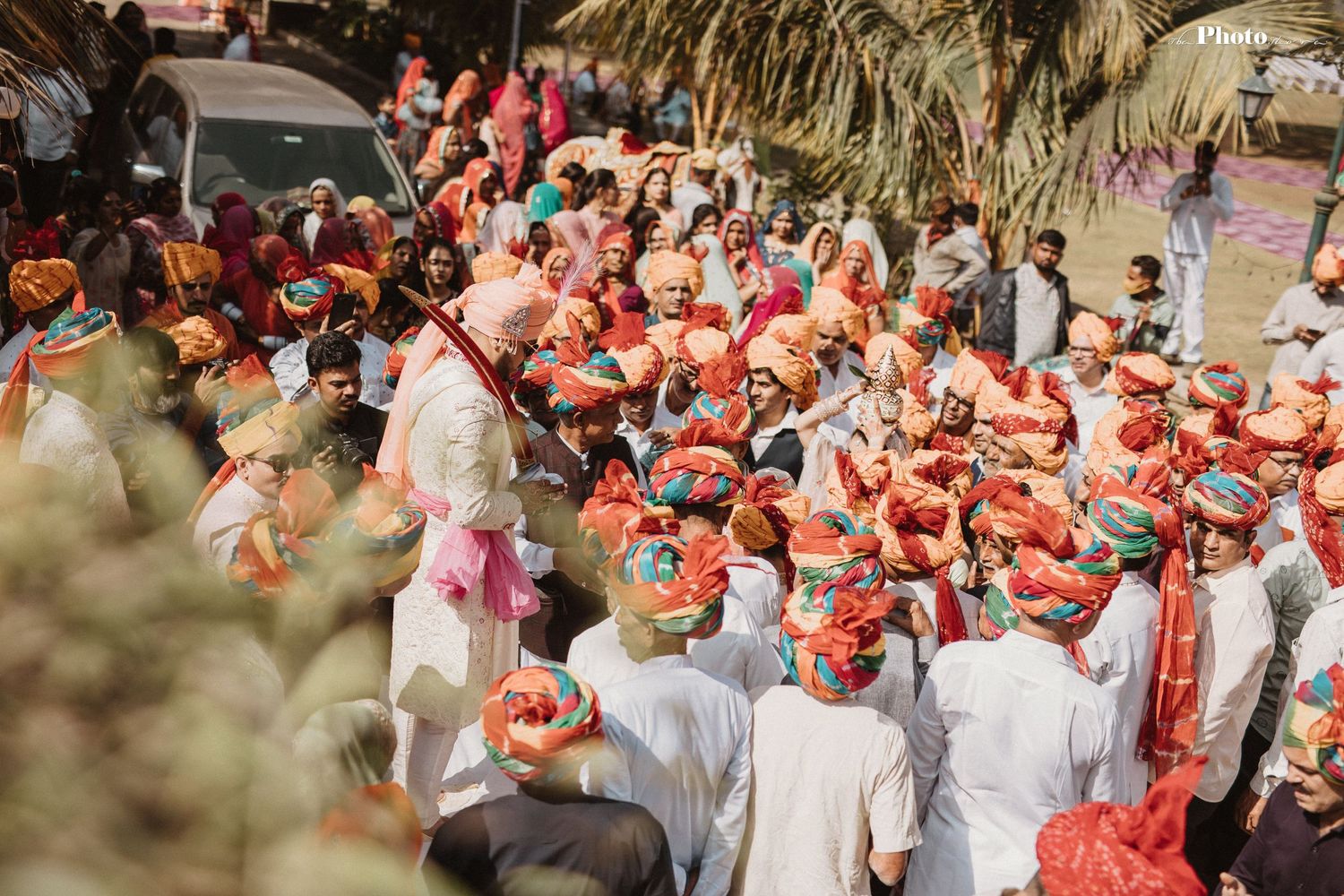 Large gathering of wedding guests in traditional Indian attire holding coral umbrellas during outdoor celebration.