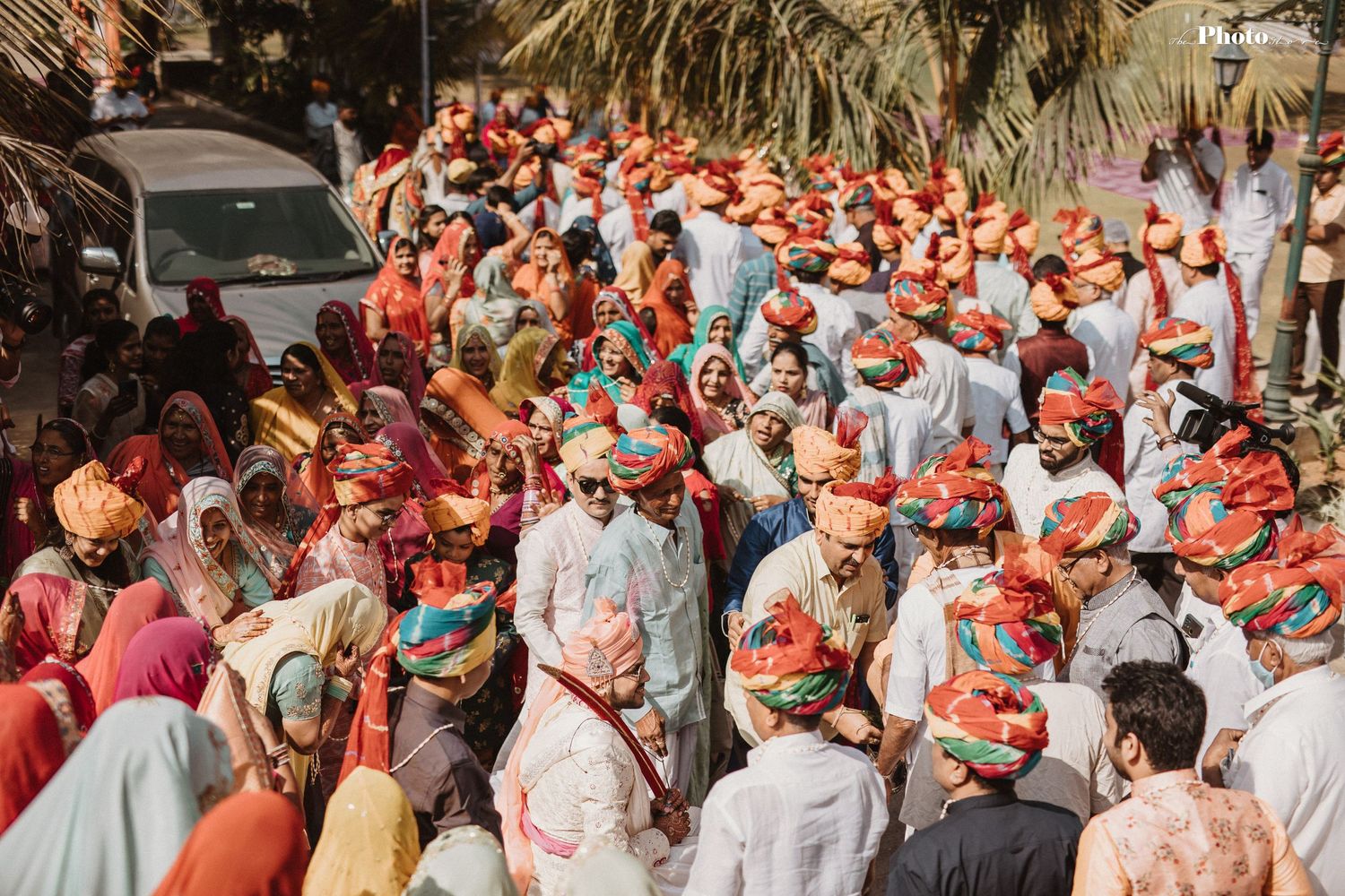 Wedding guests in traditional Indian dress hold coral parasols during an outdoor ceremony.