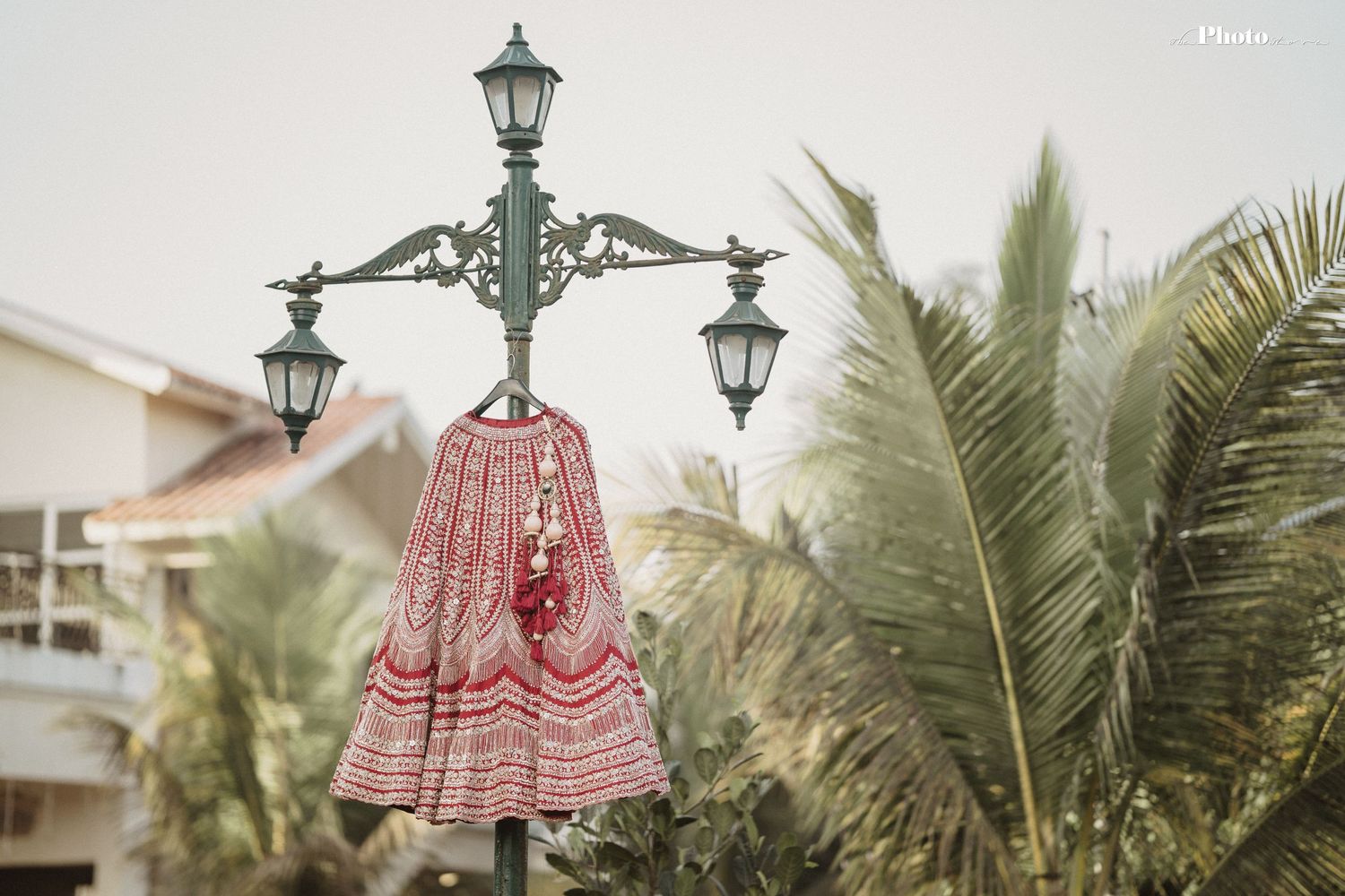 Red bridal lehenga hanging on ornate vintage lamp post with palm trees in background.