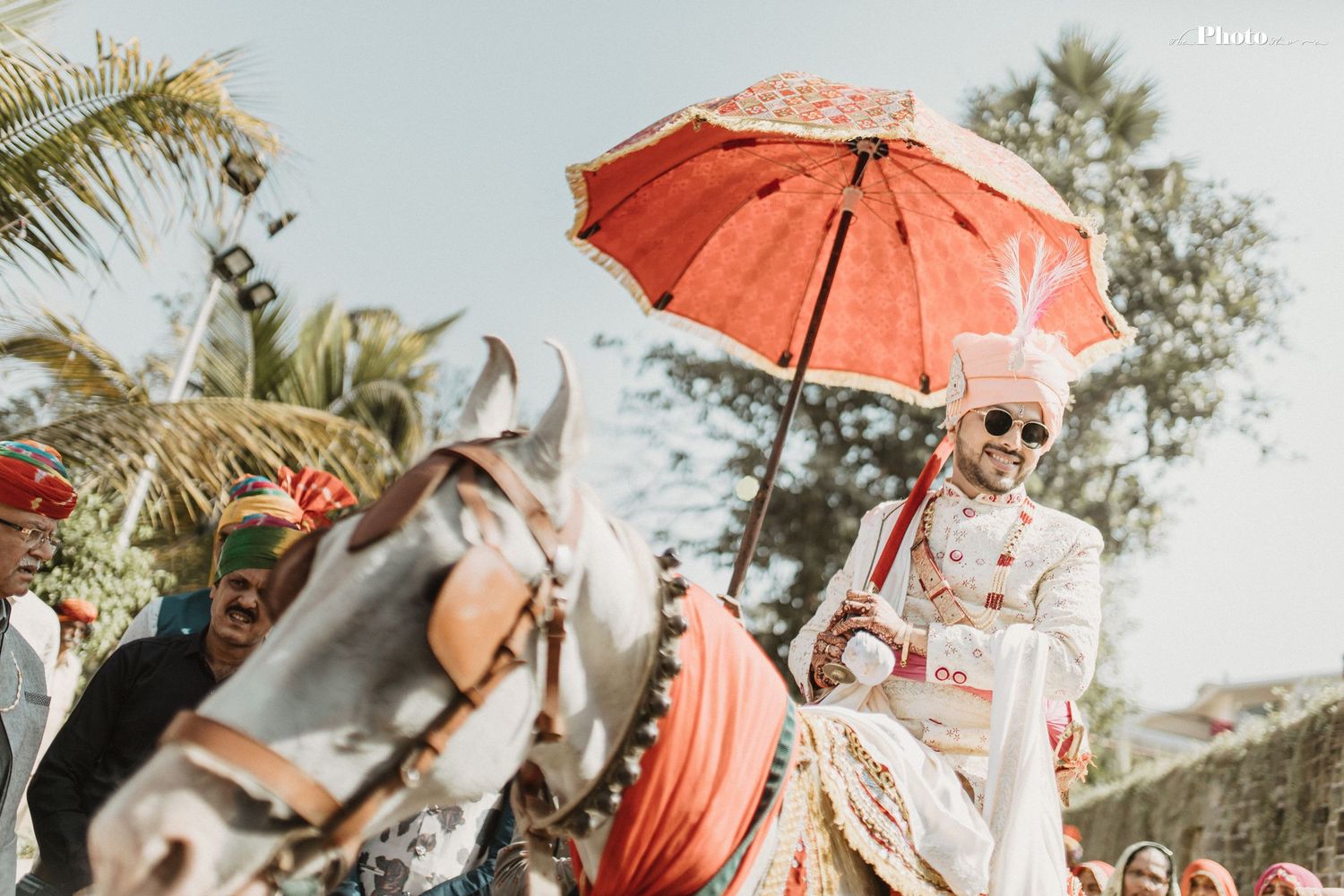 Wedding guests holding coral parasols gather under palm trees for an outdoor celebration.