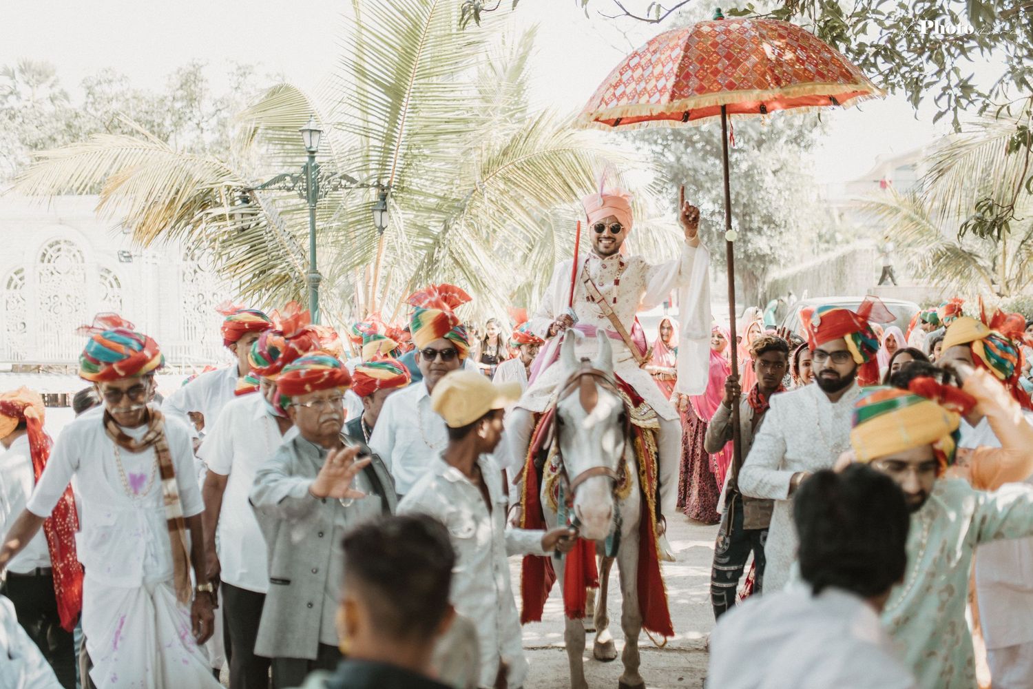 Wedding guests in traditional attire gather under coral parasols for an outdoor celebration.