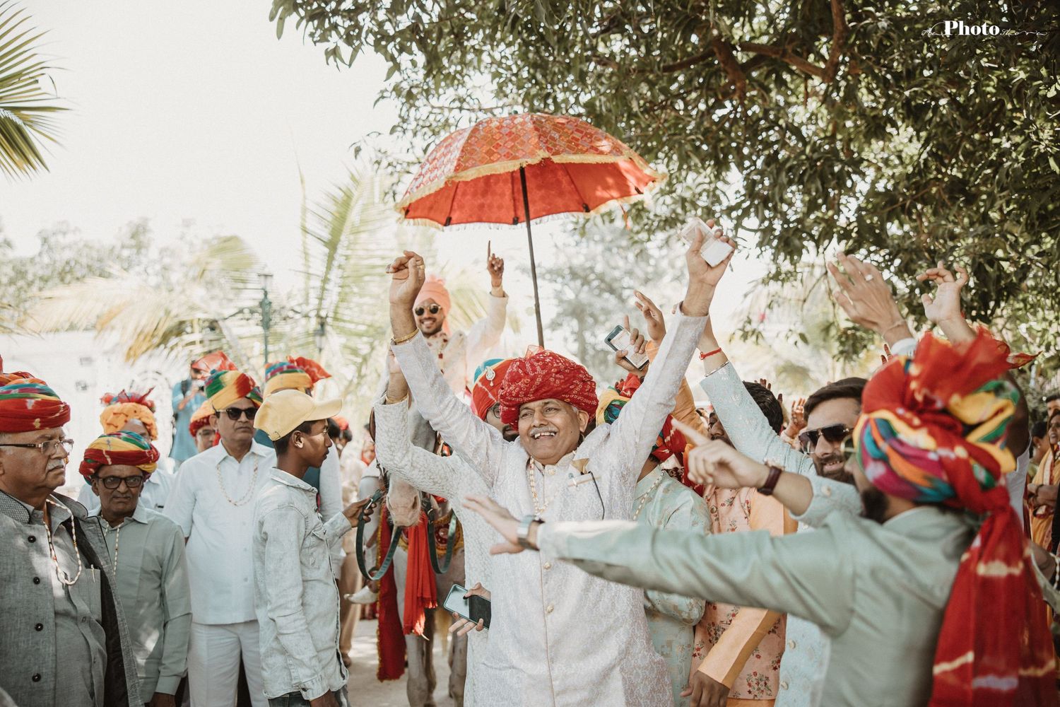 Festive crowd gathers under trees with red umbrellas during outdoor celebration.