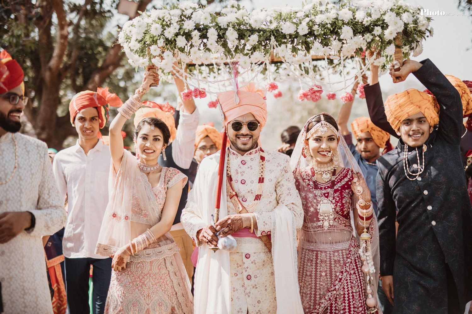 Wedding party celebrating under a floral canopy with pink and white decorations at an outdoor Indian ceremony.
