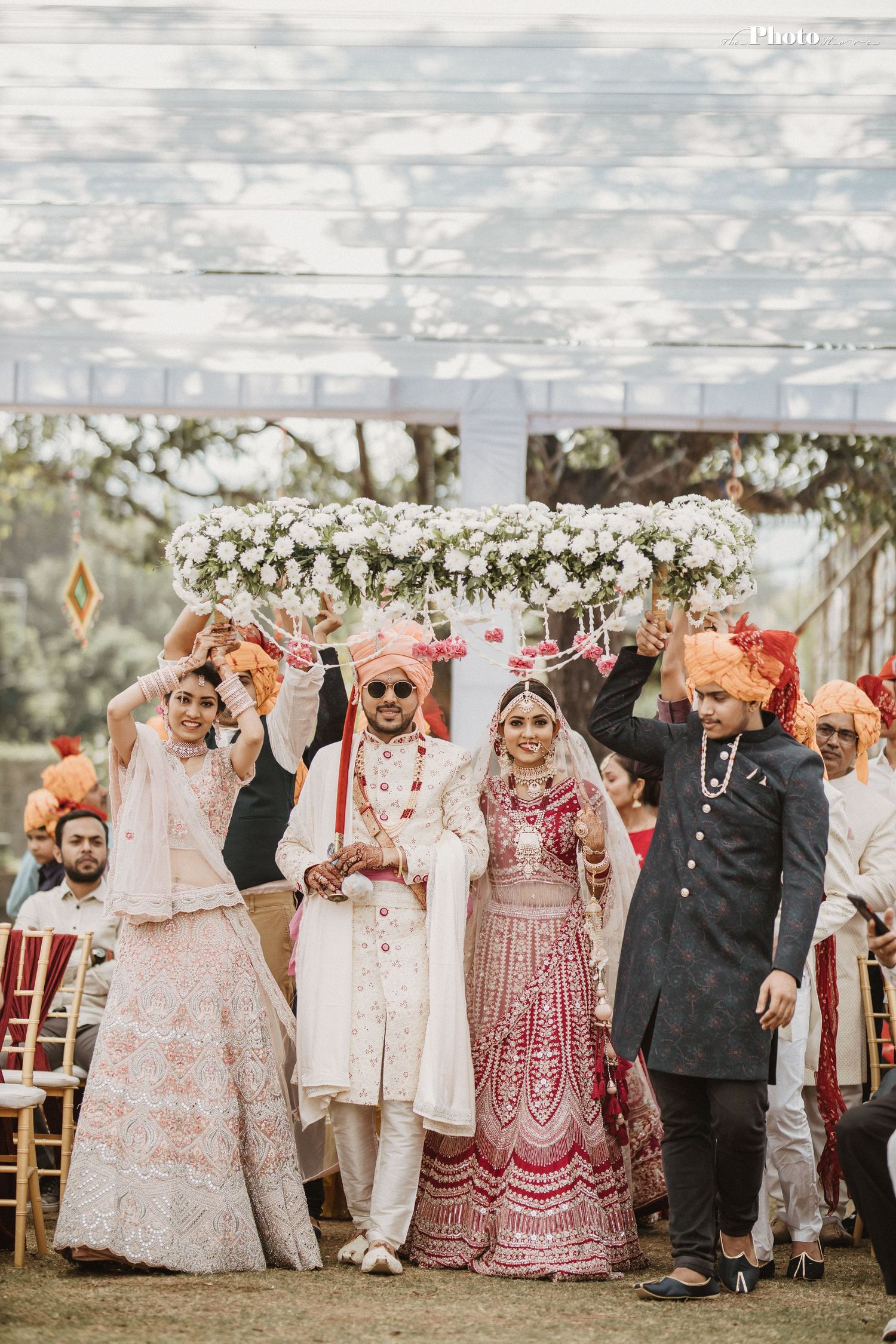 Wedding group photo under white floral arch with bride in red lehenga and groom in white traditional attire.