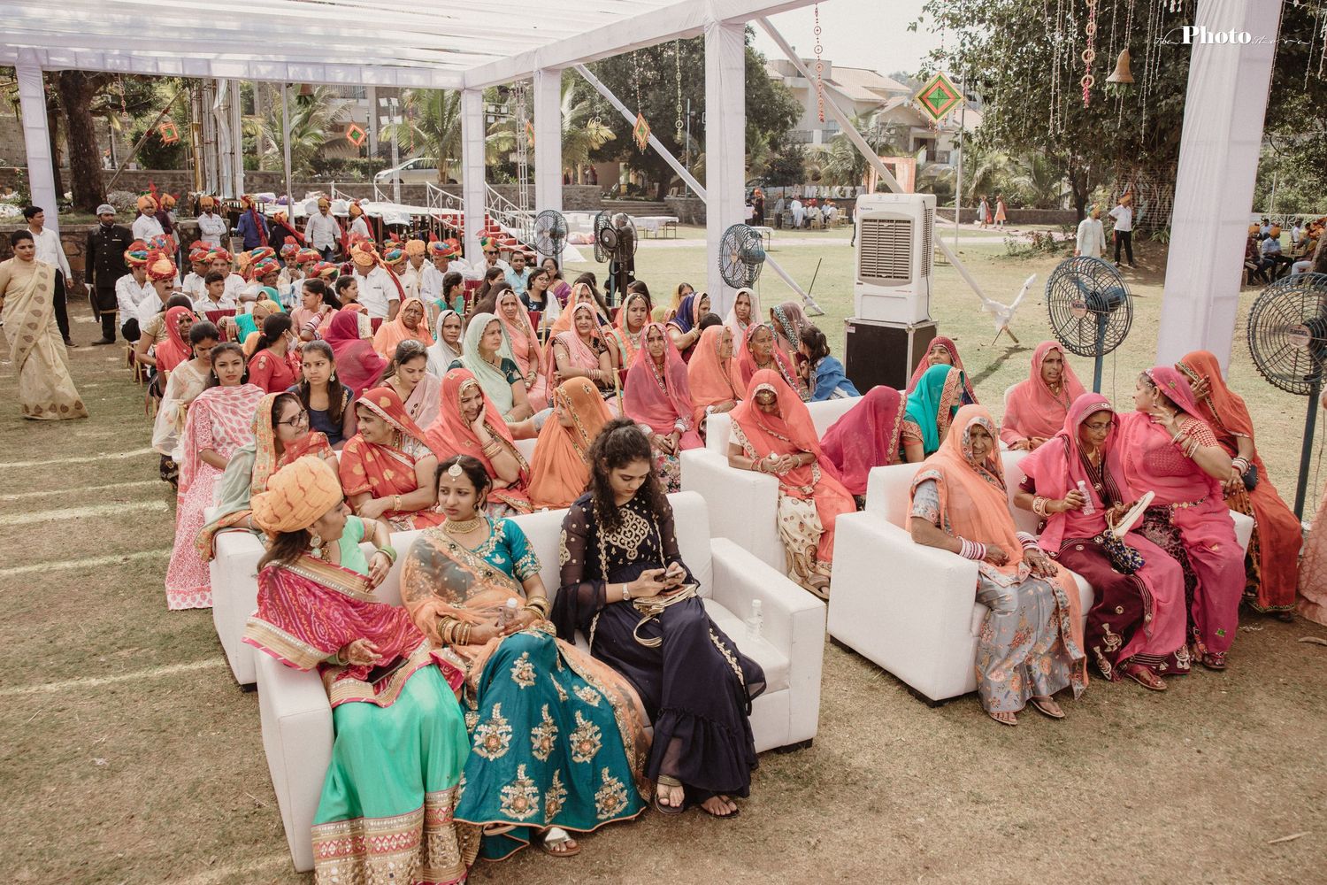 Wide view of wedding guests seated under white canopy during an outdoor Indian celebration.