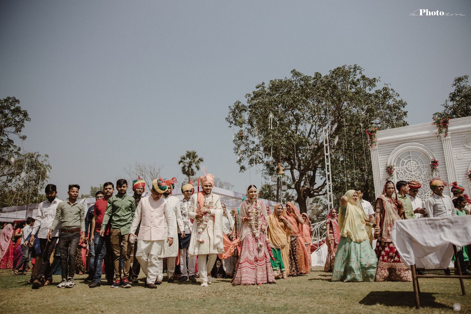 Large wedding party poses together on grassy lawn during daytime celebration.