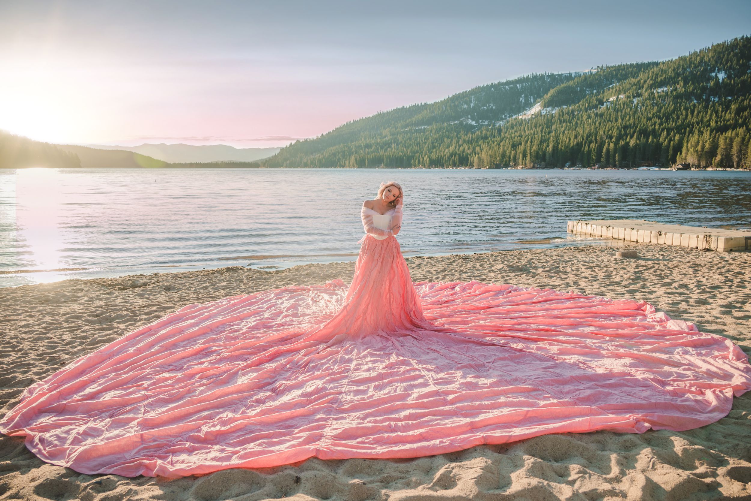Woman in a flowing pink parachute gown with a long train standing on a sandy lakeshore at sunset.
