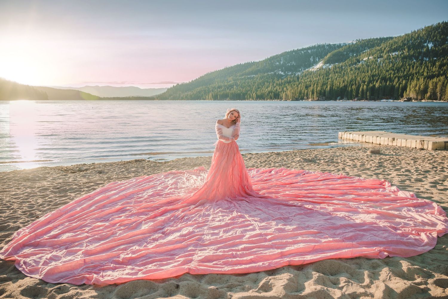Woman in a flowing pink parachute gown with a long train standing on a sandy lakeshore at sunset.