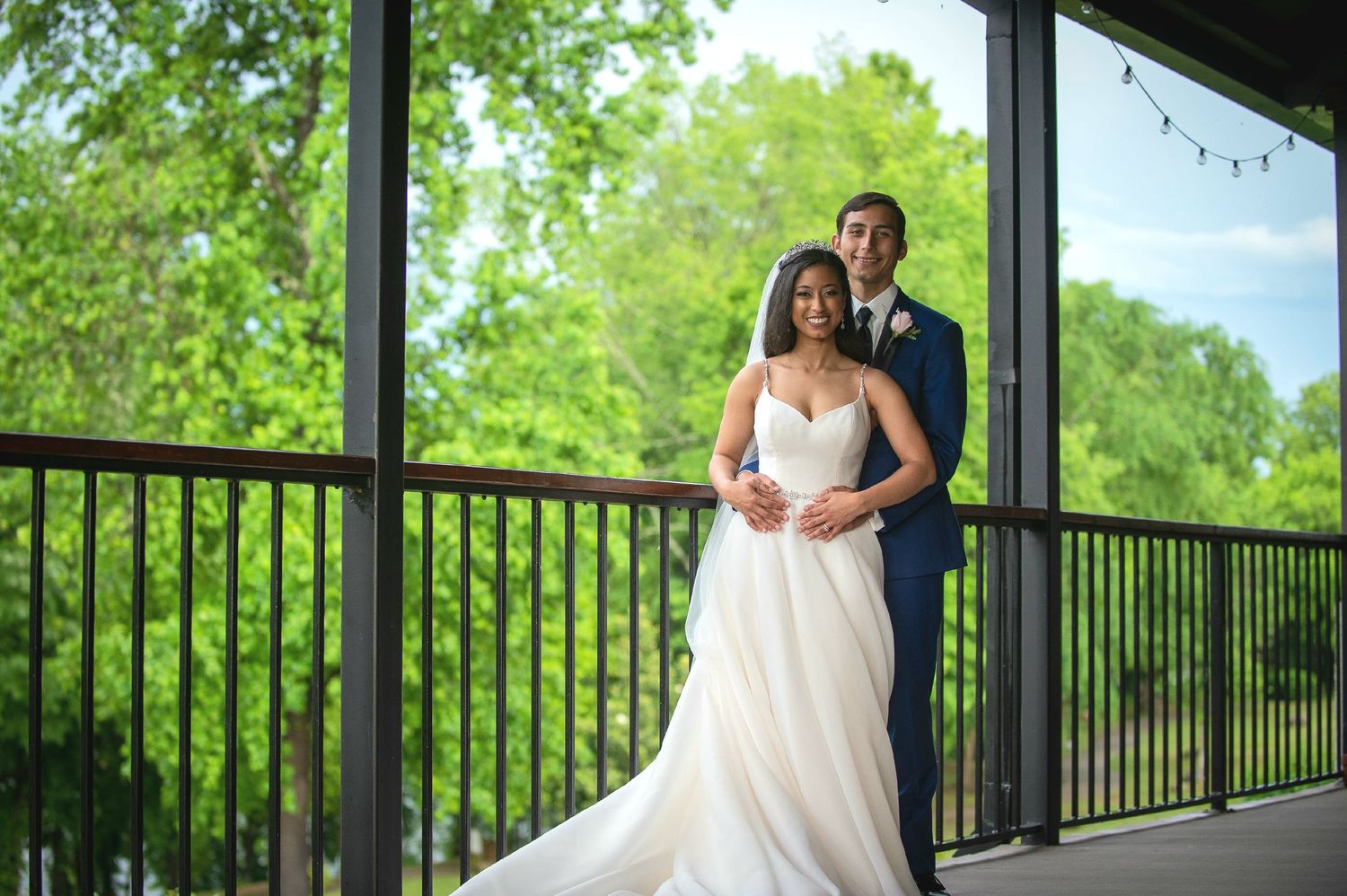 A romantic wedding photo of a couple embracing on a covered deck overlooking greenery.