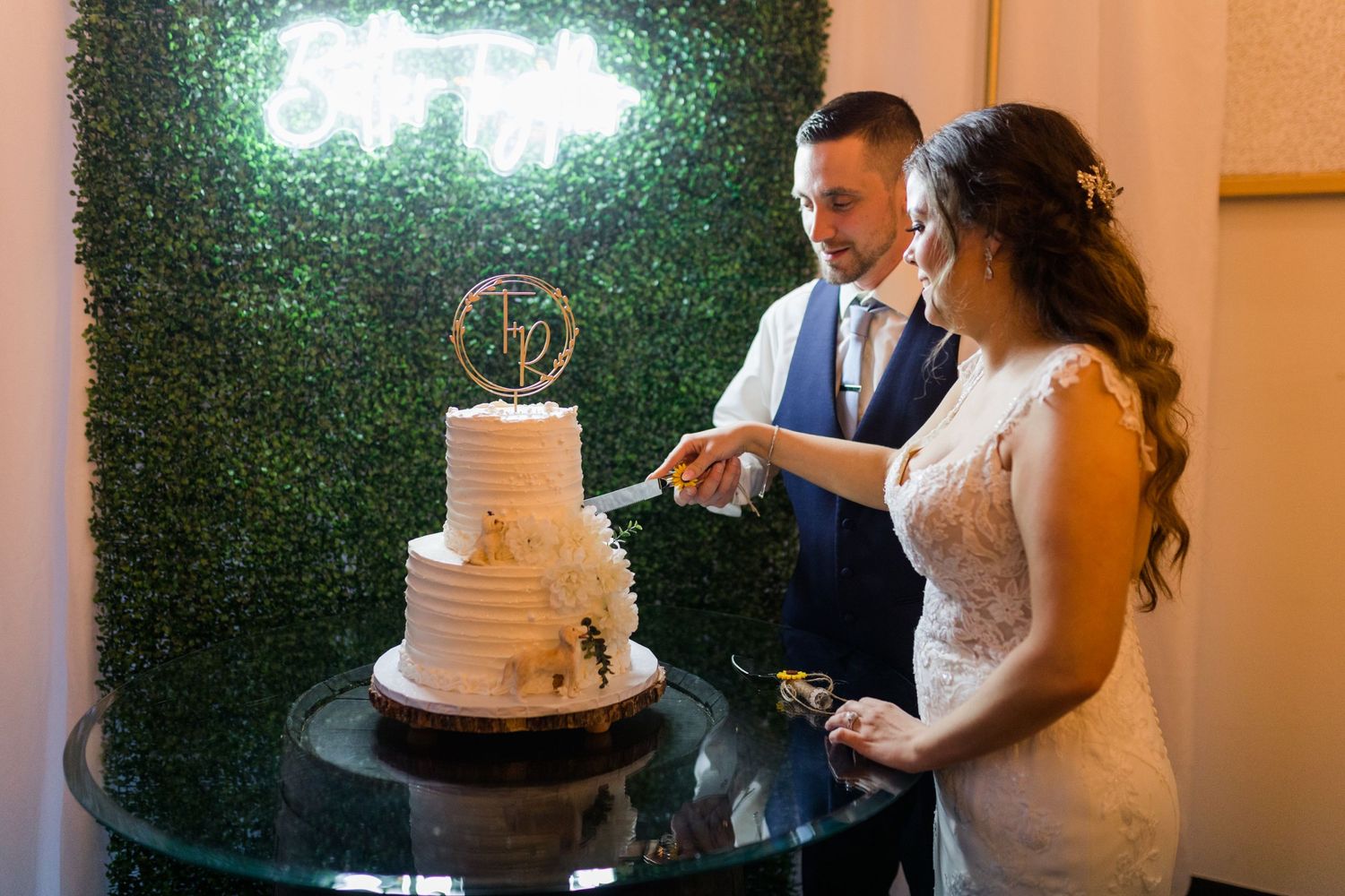 Wedding couple cutting their cake together against green backdrop with guests watching.