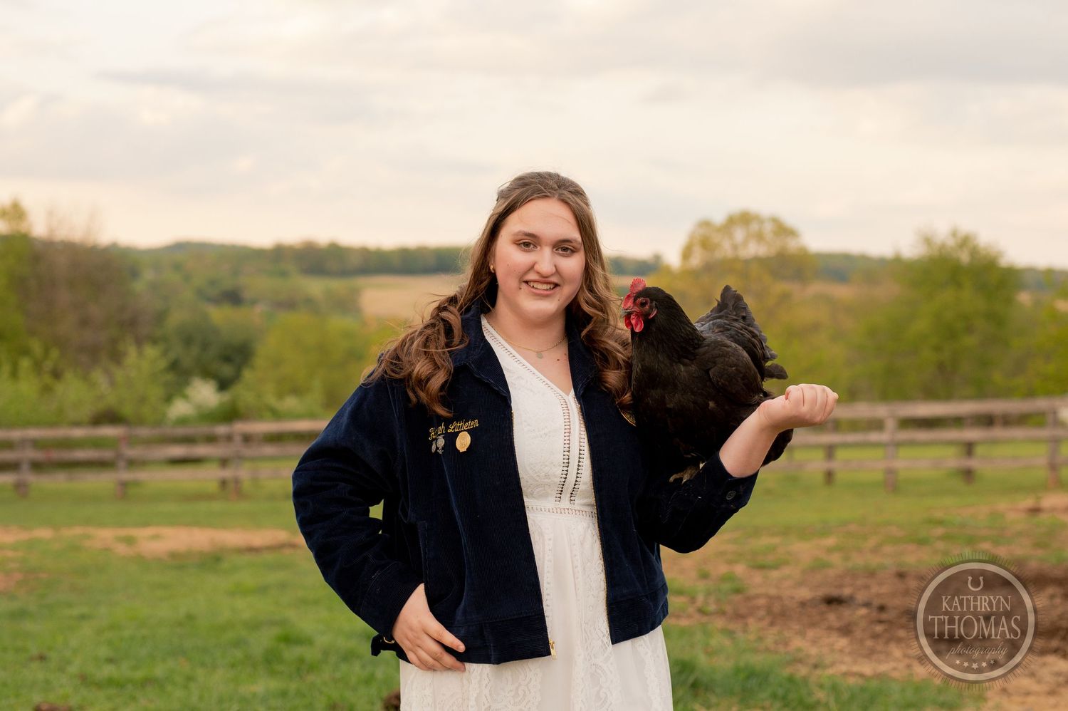 A white-dressed person in navy jacket poses outdoors with a black chicken on rolling farmland.