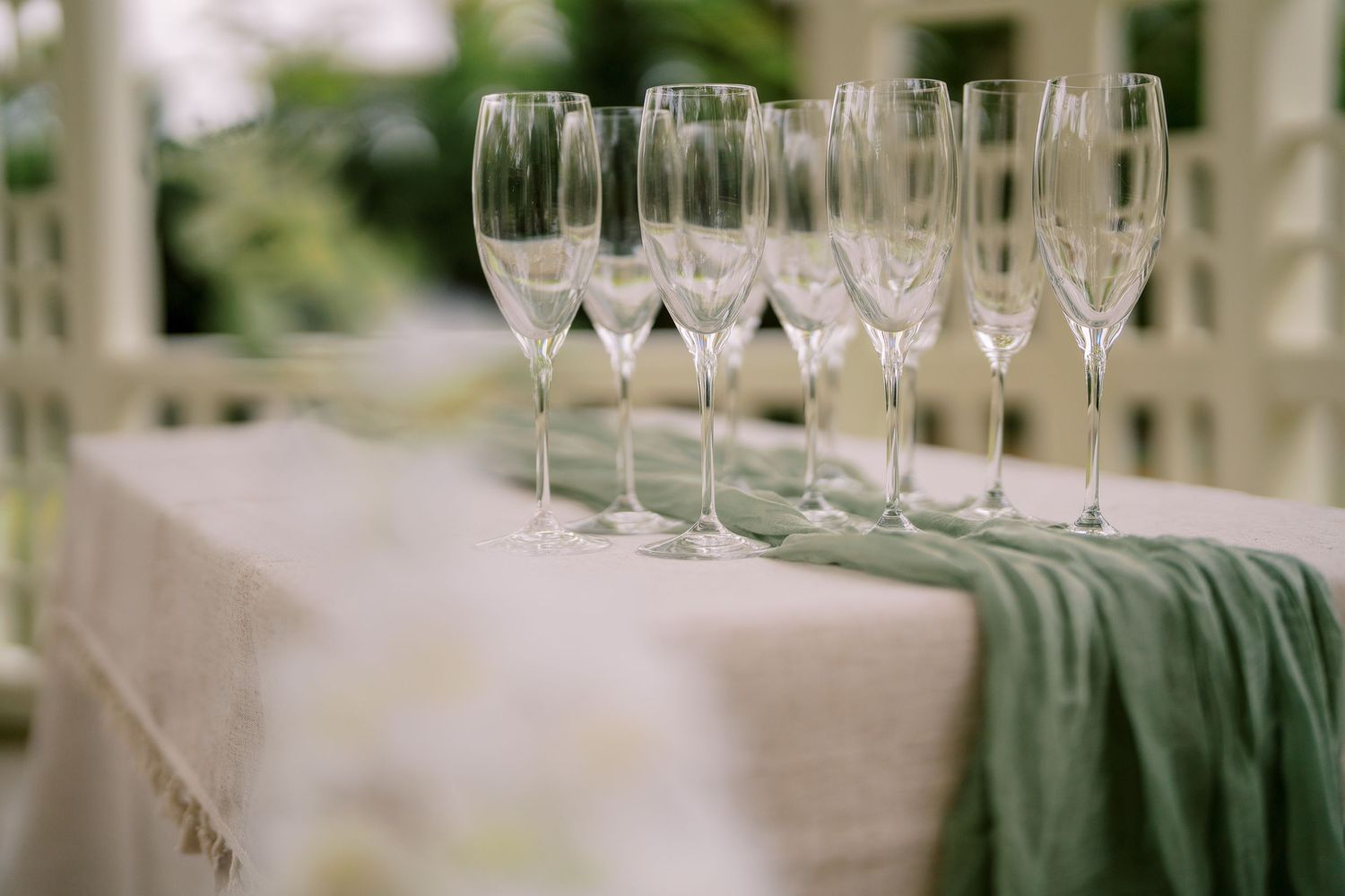 Row of empty champagne flutes arranged on a table with white tablecloth and sage green runner.