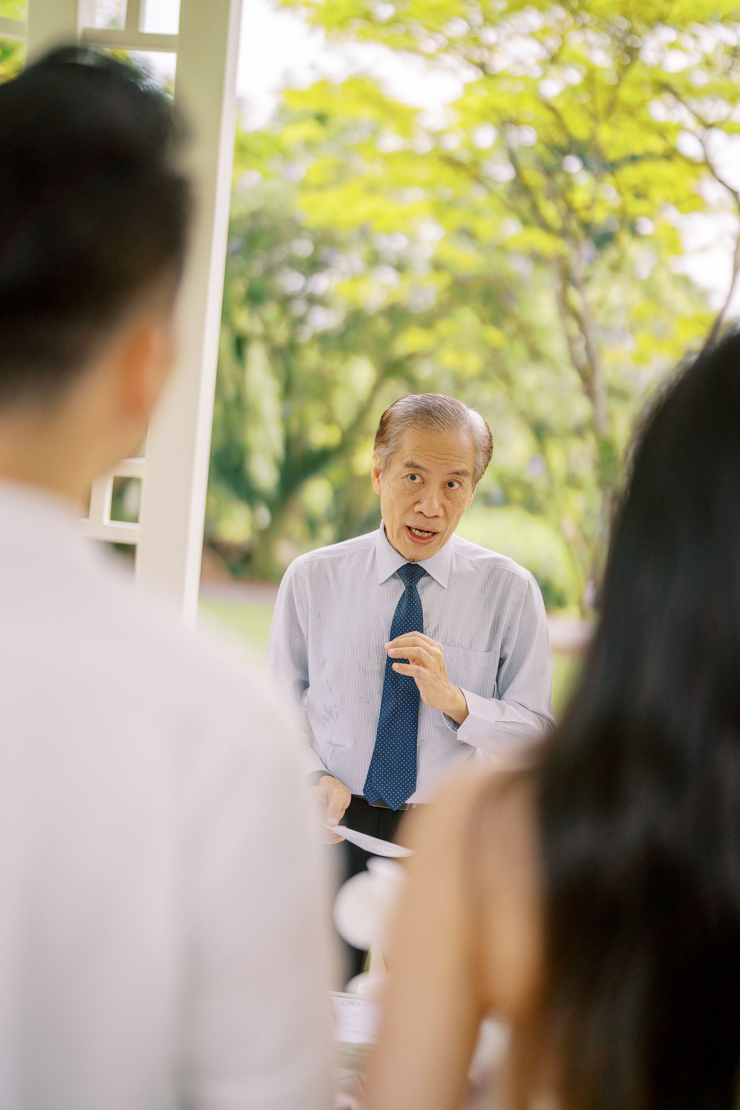 A speaker in formal attire presents to an audience in an outdoor garden setting.