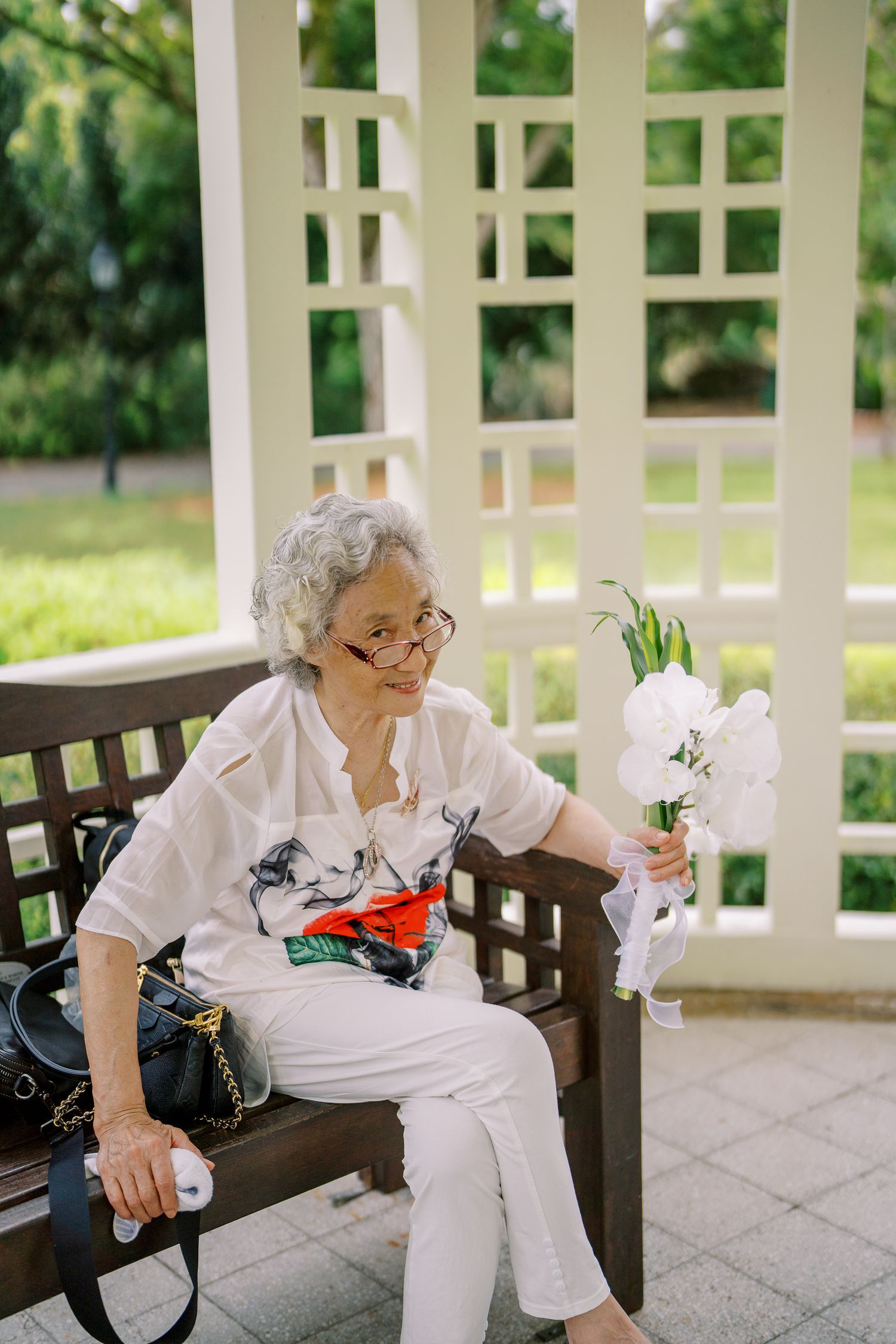 Person sitting on wooden bench in white gazebo with flowers.