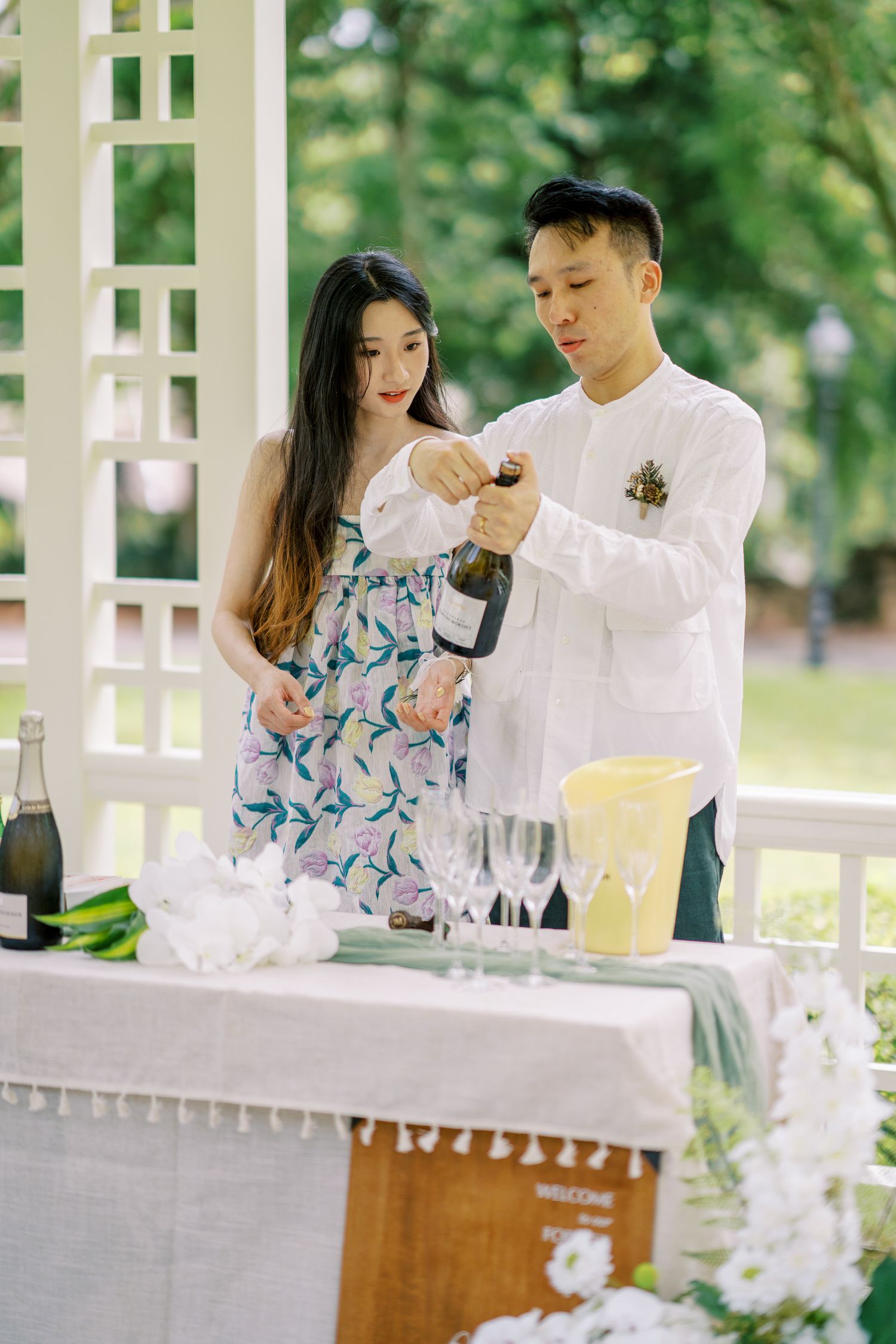 Couple pouring champagne at an outdoor celebration table decorated with white flowers.