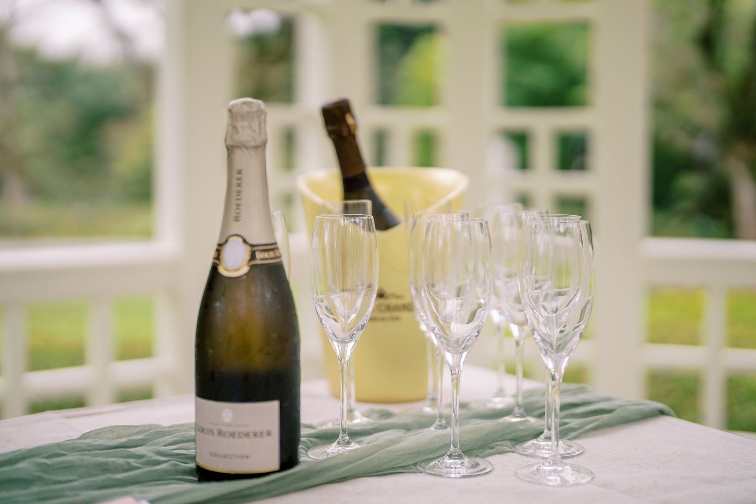 Champagne bottle and glasses arranged on table with green runner in outdoor setting.